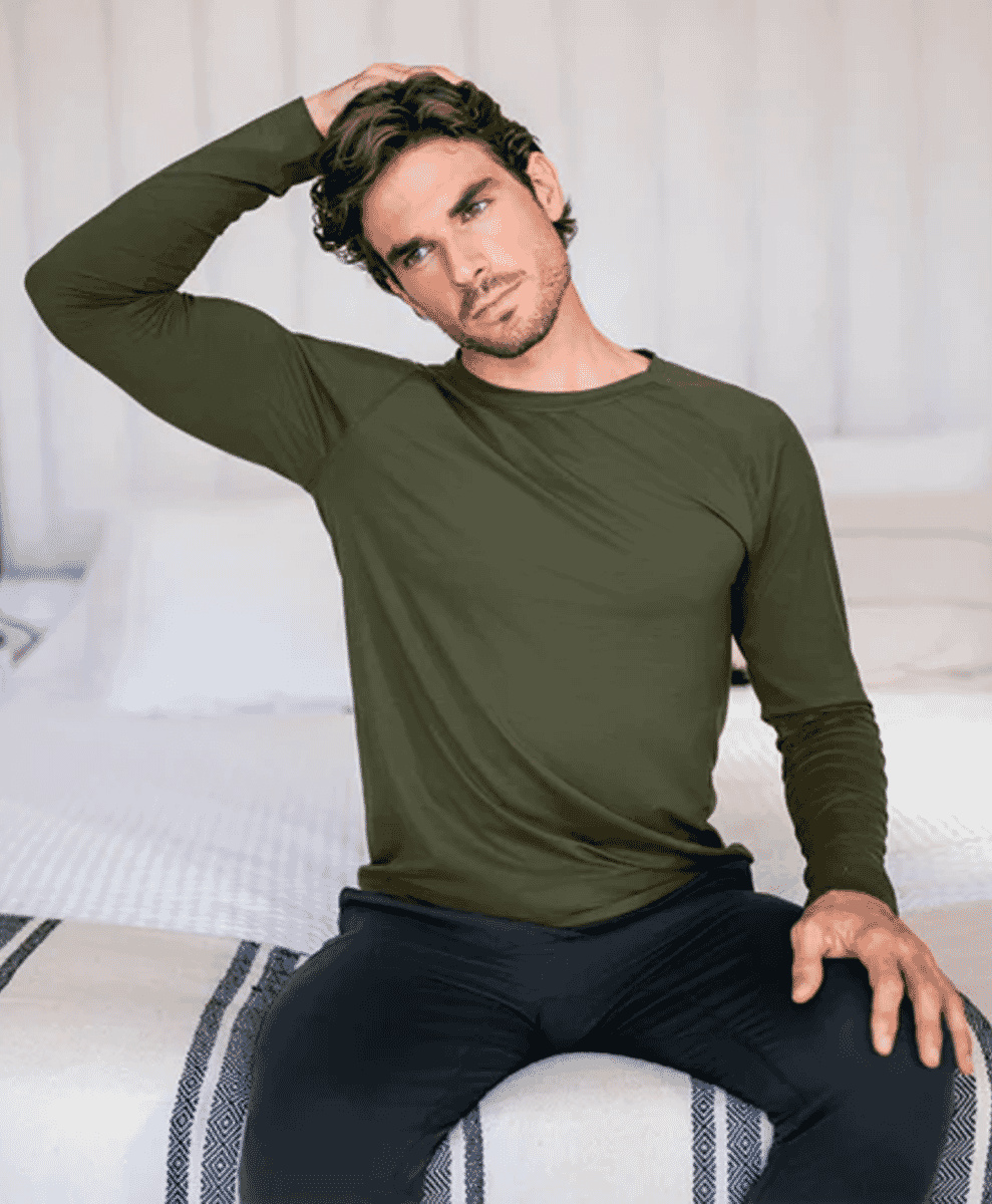 Man in a fitted olive green long-sleeve top and black pants sitting on a bed.