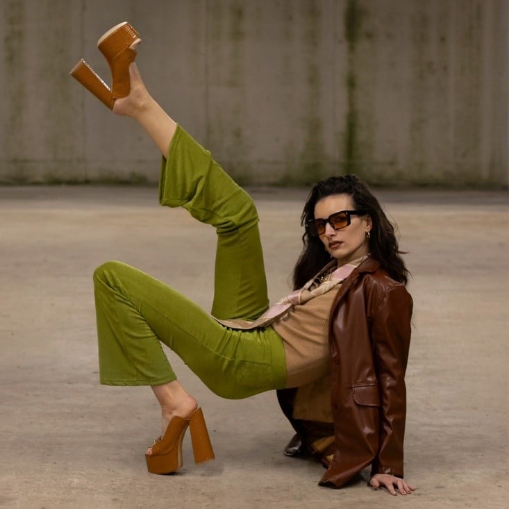 A model in olive green pants and a leather jacket wearing tan platform mules in a gritty urban setting.