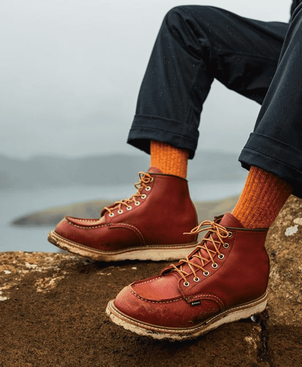 Person sitting on a rocky cliff edge wearing red leather work boots and orange socks with a misty ocean background.