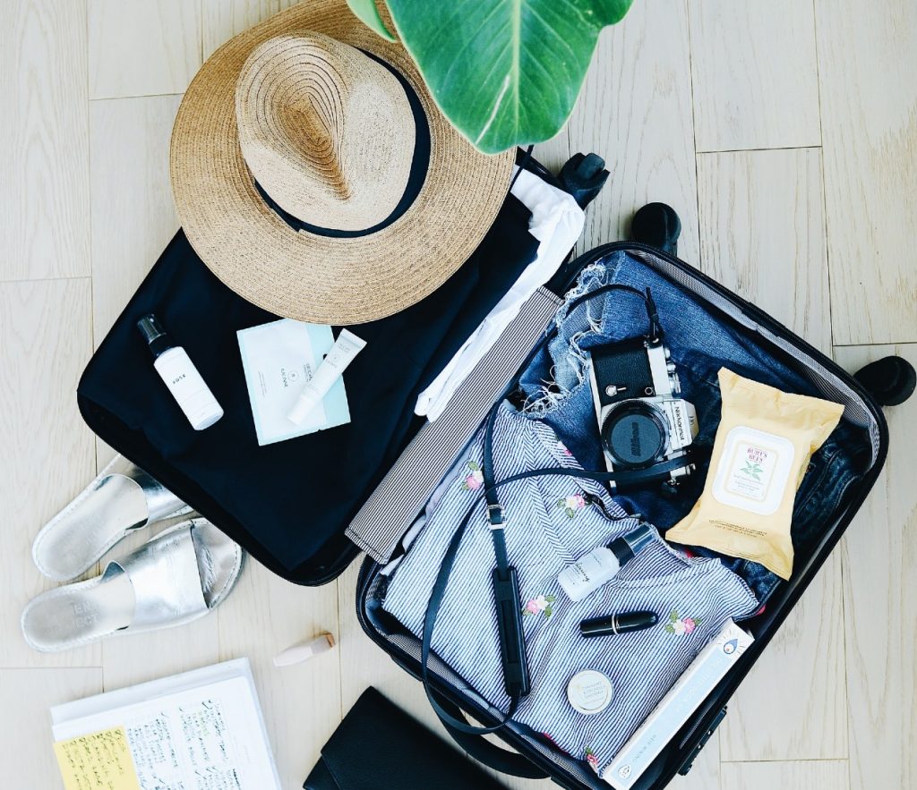 Overhead view of a light wood floor with an open suitcase containing neatly packed summer clothes, a camera, and a straw hat.