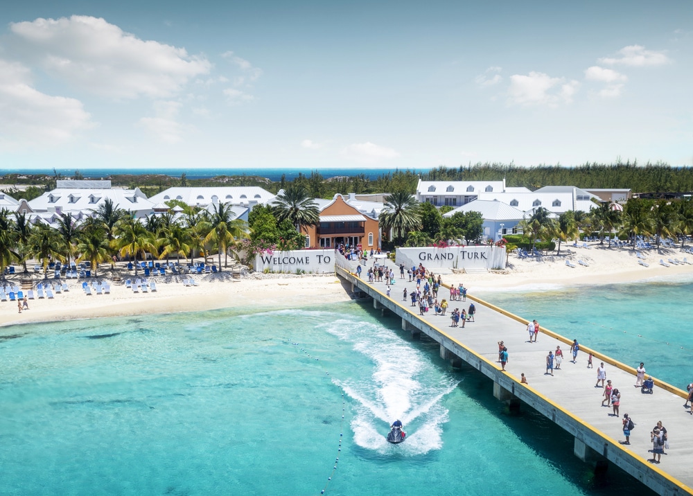 People in breezy resort attire walking along a sunny pier by turquoise ocean waters.