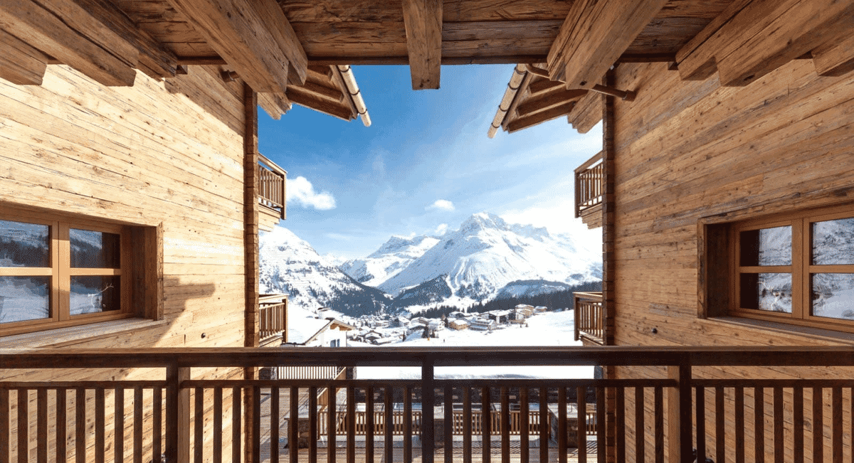 Winter vista of snow-capped peaks seen from a rustic wooden chalet balcony under a blue sky.