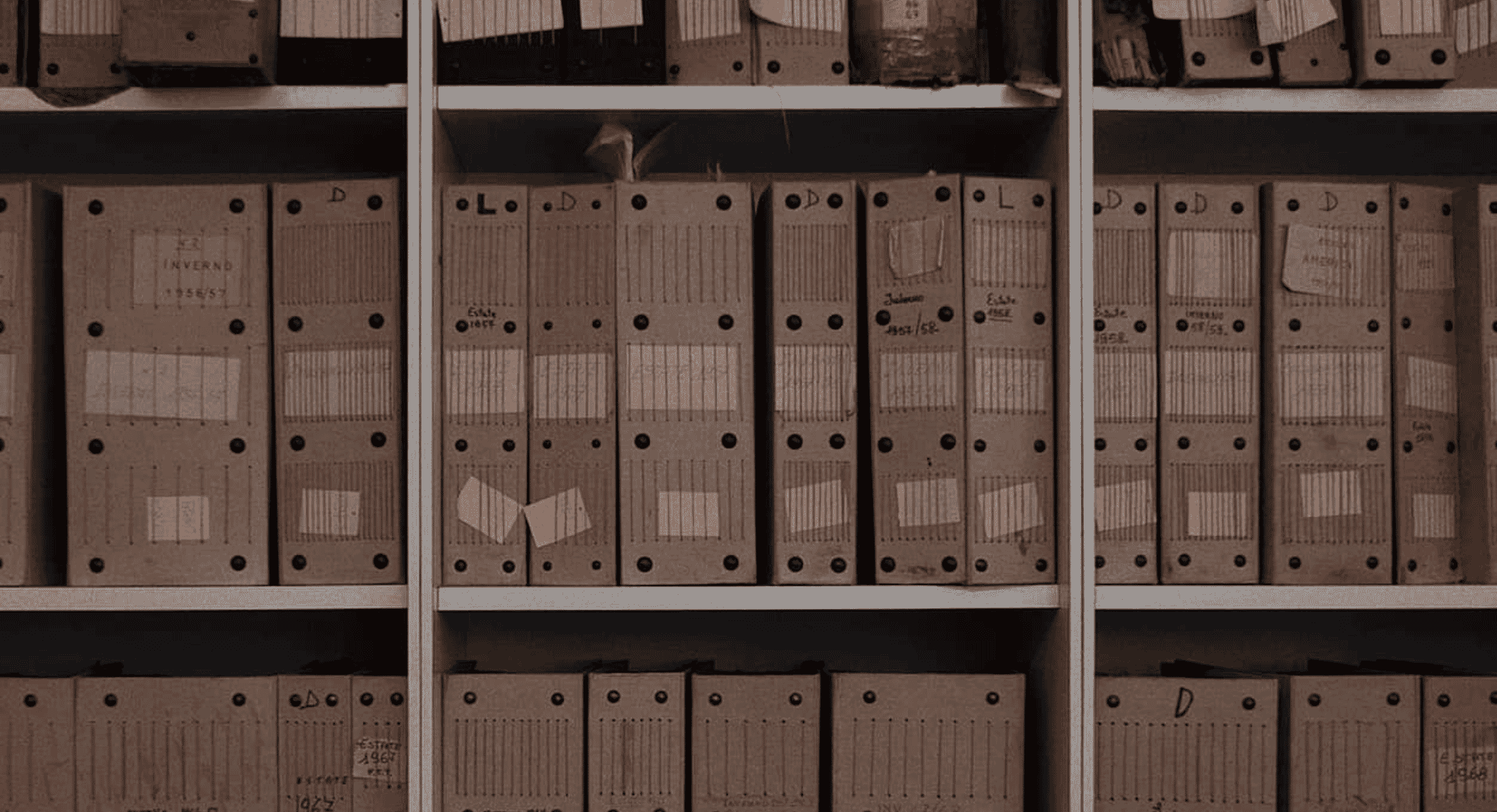 Rows of vintage cardboard file folders with handwritten labels on library shelves.