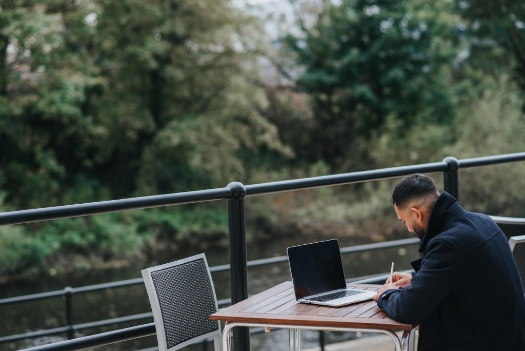 A man in a dark coat sitting at an outdoor wooden table with a laptop overlooking greenery.