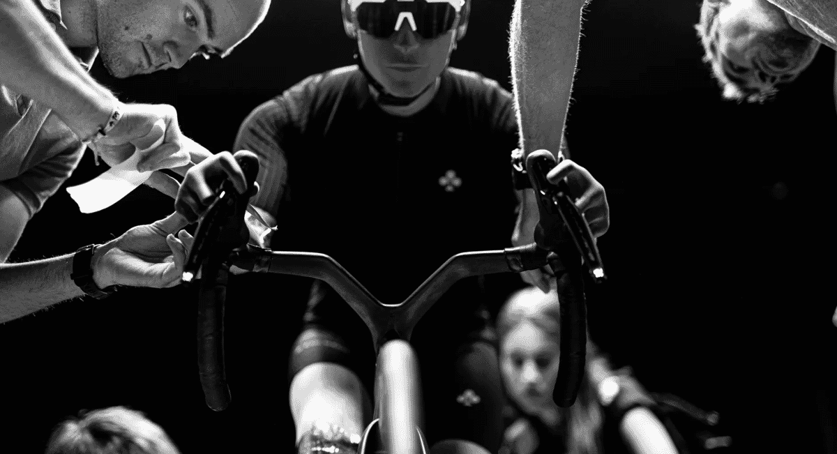 Black and white photo of a cyclist in an aerodynamic jersey being adjusted on a stationary bike by team members.