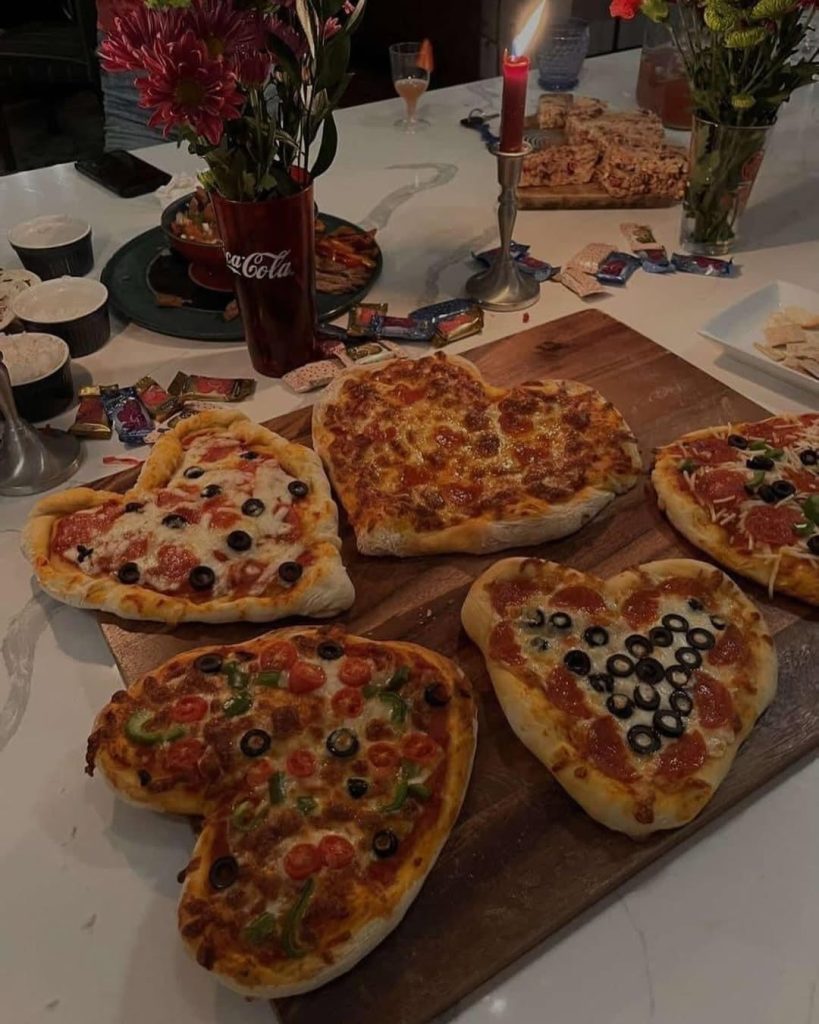 Heart-shaped pizzas on a wooden serving board surrounded by candles and flowers.