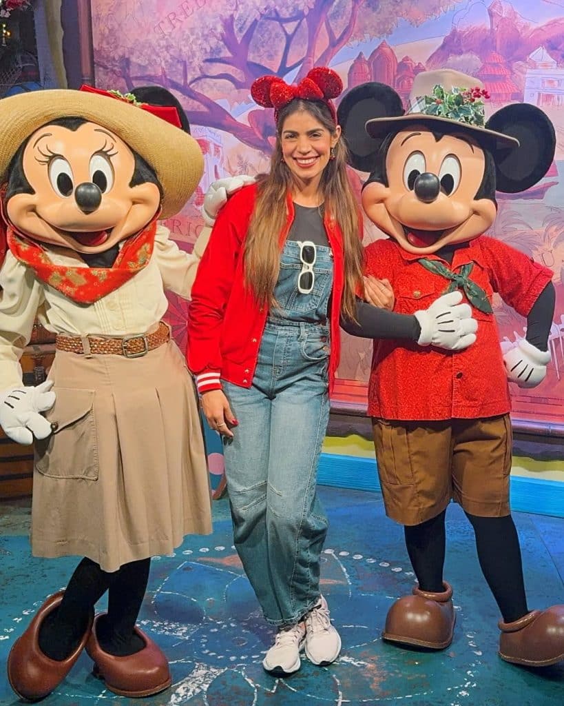 A woman in a red bomber jacket and denim overalls posing with Safari-themed Mickey and Minnie characters.