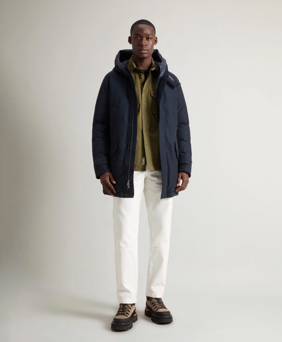 Man modeling a navy blue Woolrich hooded puffer coat with white trousers and boots.