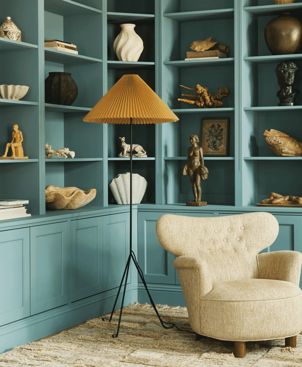 A distinctive pleated floor lamp with a warm shade standing in front of teal built-in bookshelves and a beige armchair.