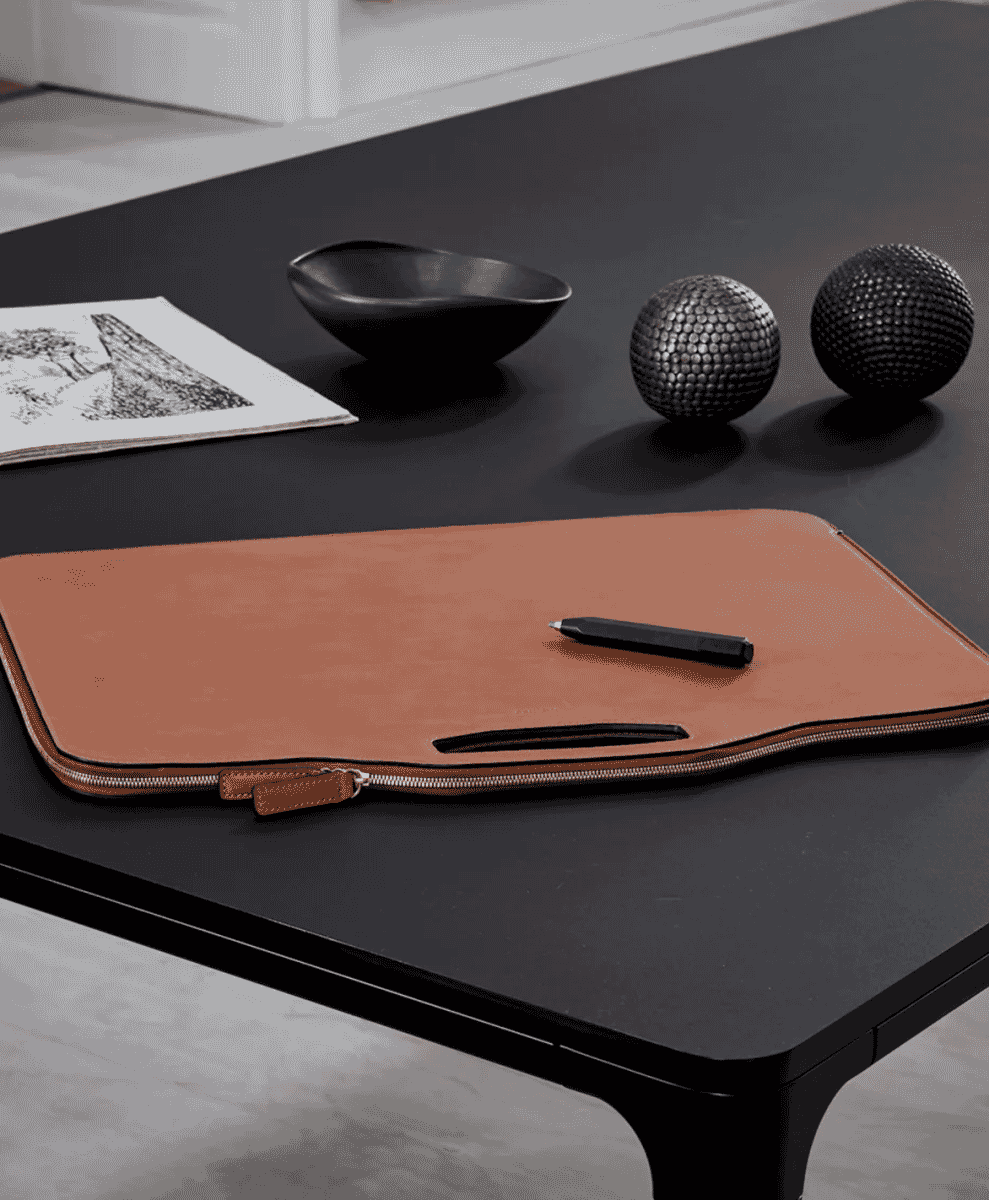 Tan leather laptop sleeve with a handle resting on a minimalist black desk with office accessories.