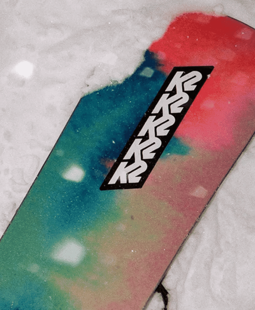 A colorful K2 snowboard with a red and teal gradient design resting on snow.