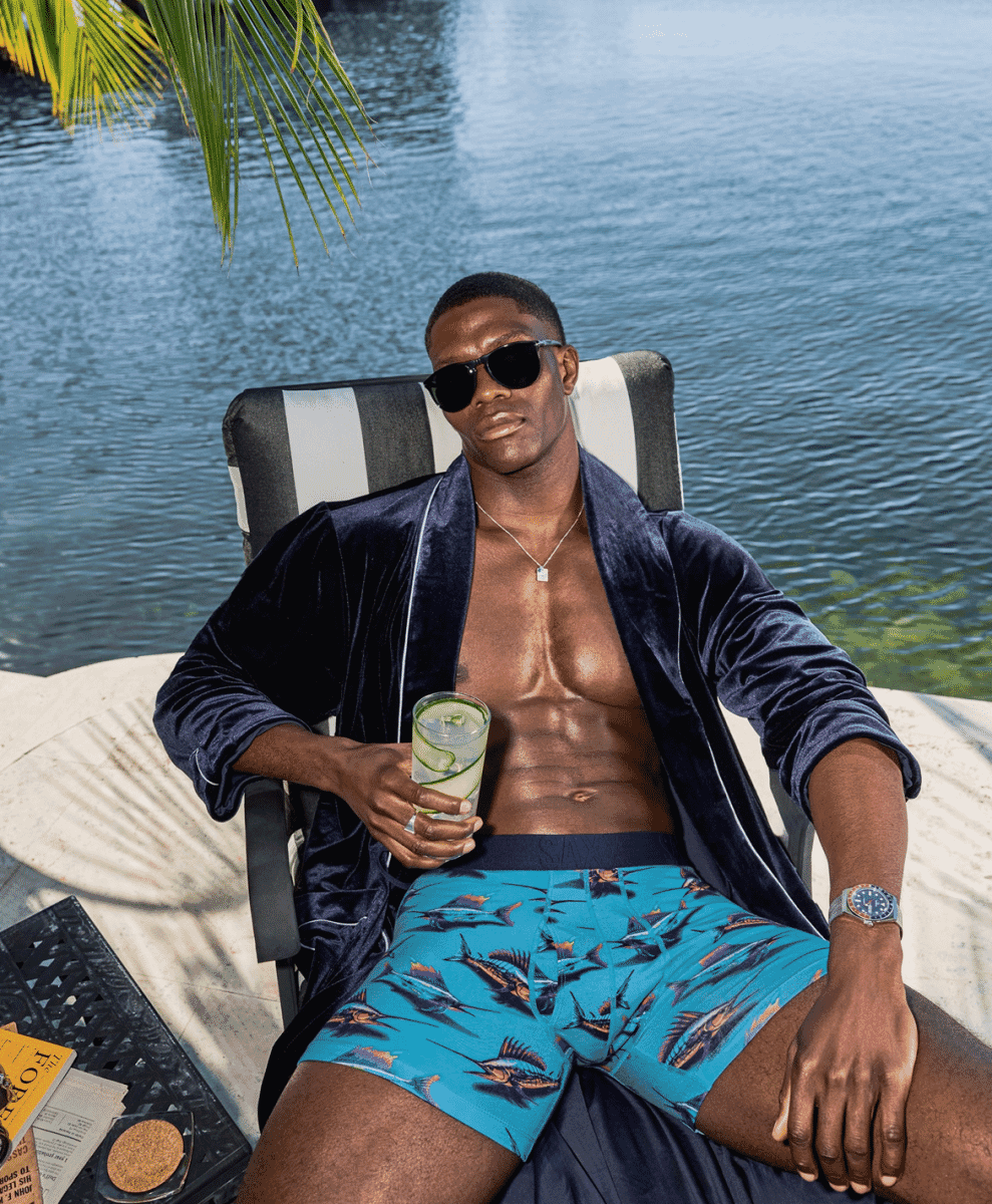 A man in a navy velvet robe and fish-print boxers lounging by a pool with a cocktail.