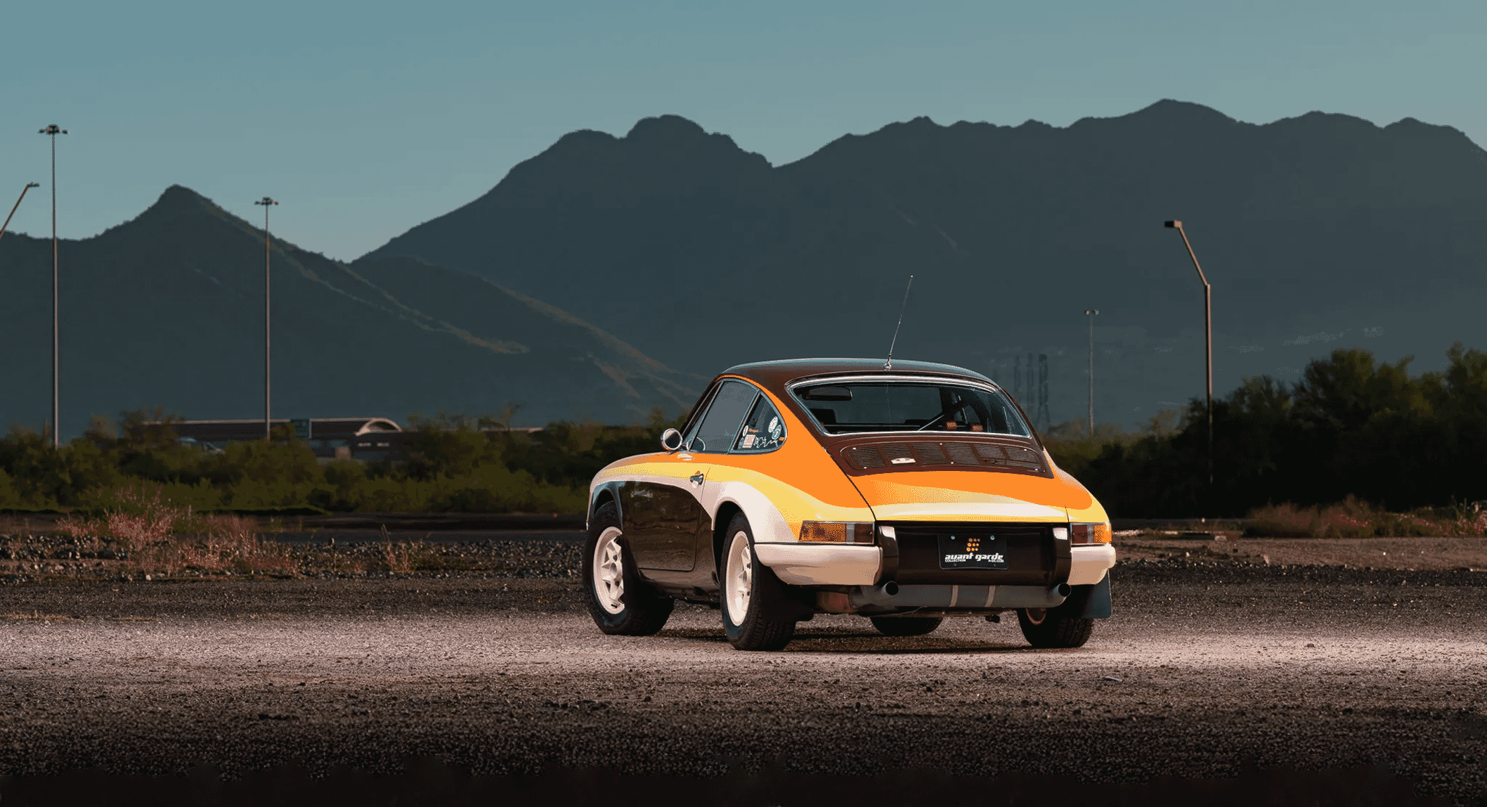 A vintage orange and black Porsche rally car parked on gravel with mountains in the background.