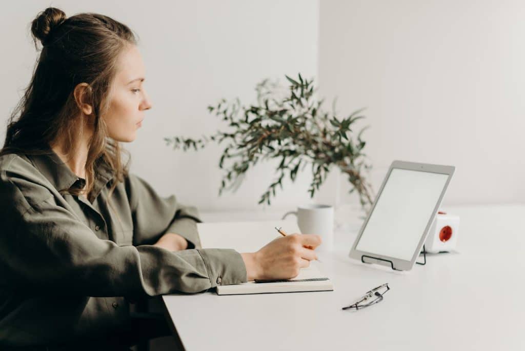 Woman in an olive-green button-up shirt writing in a notebook at a minimalist white desk with a plant.