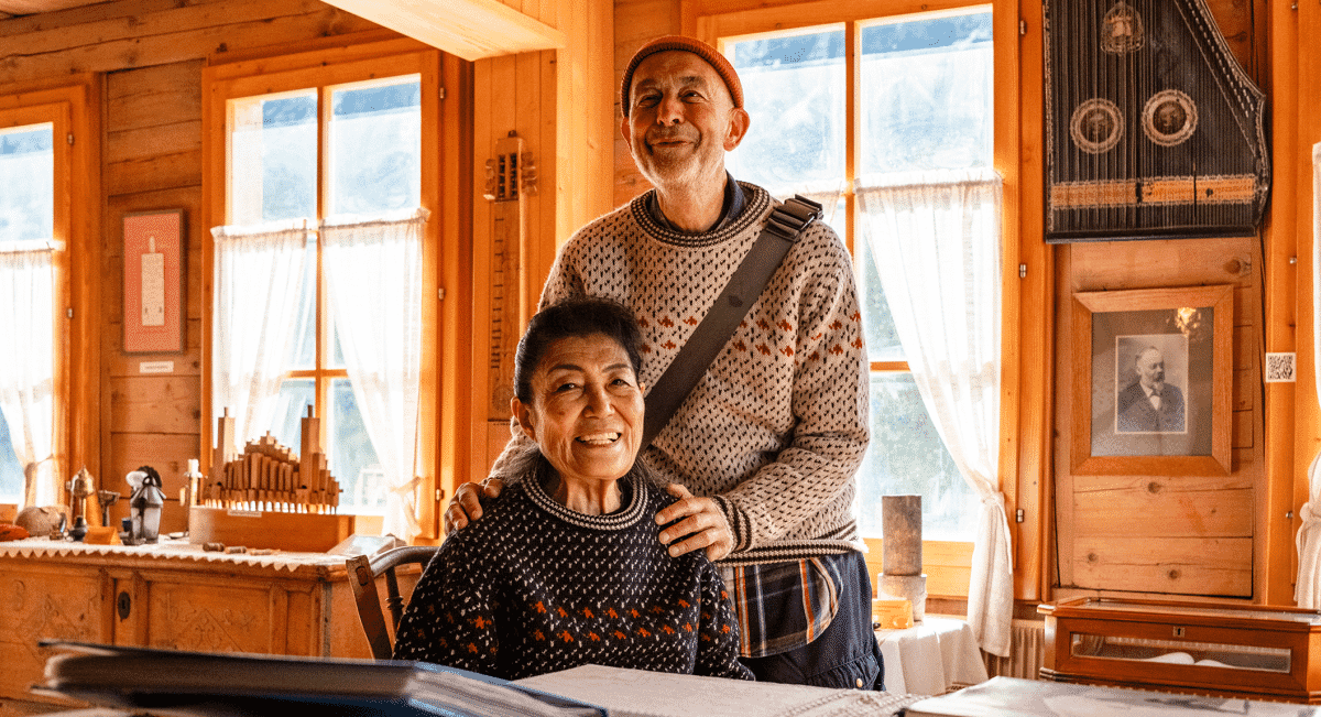An elderly man and woman in heritage knitwear sitting inside a vintage wooden cabin.