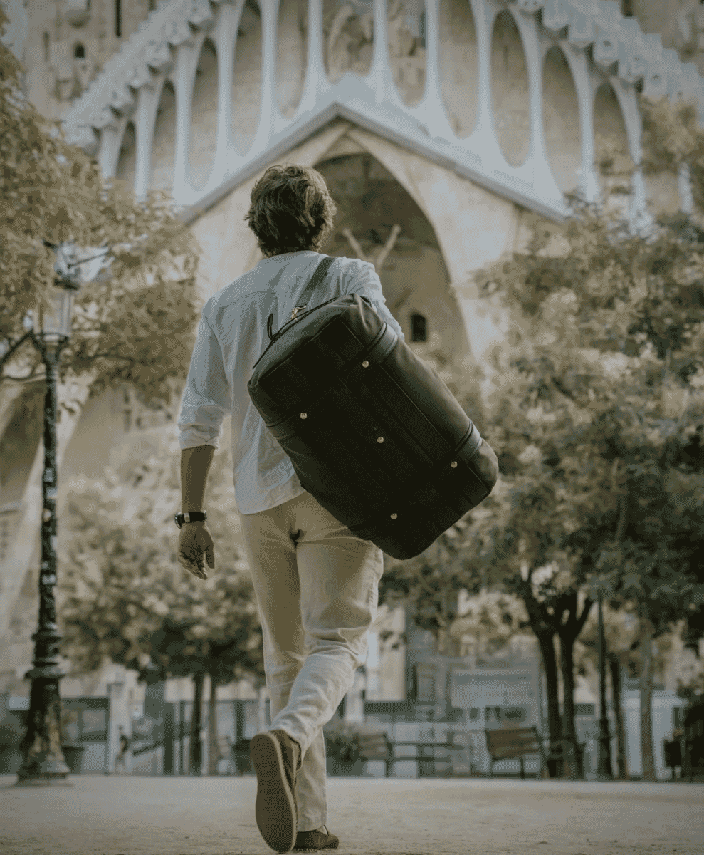 Man carrying a black leather duffel bag against a backdrop of classic architecture and trees.