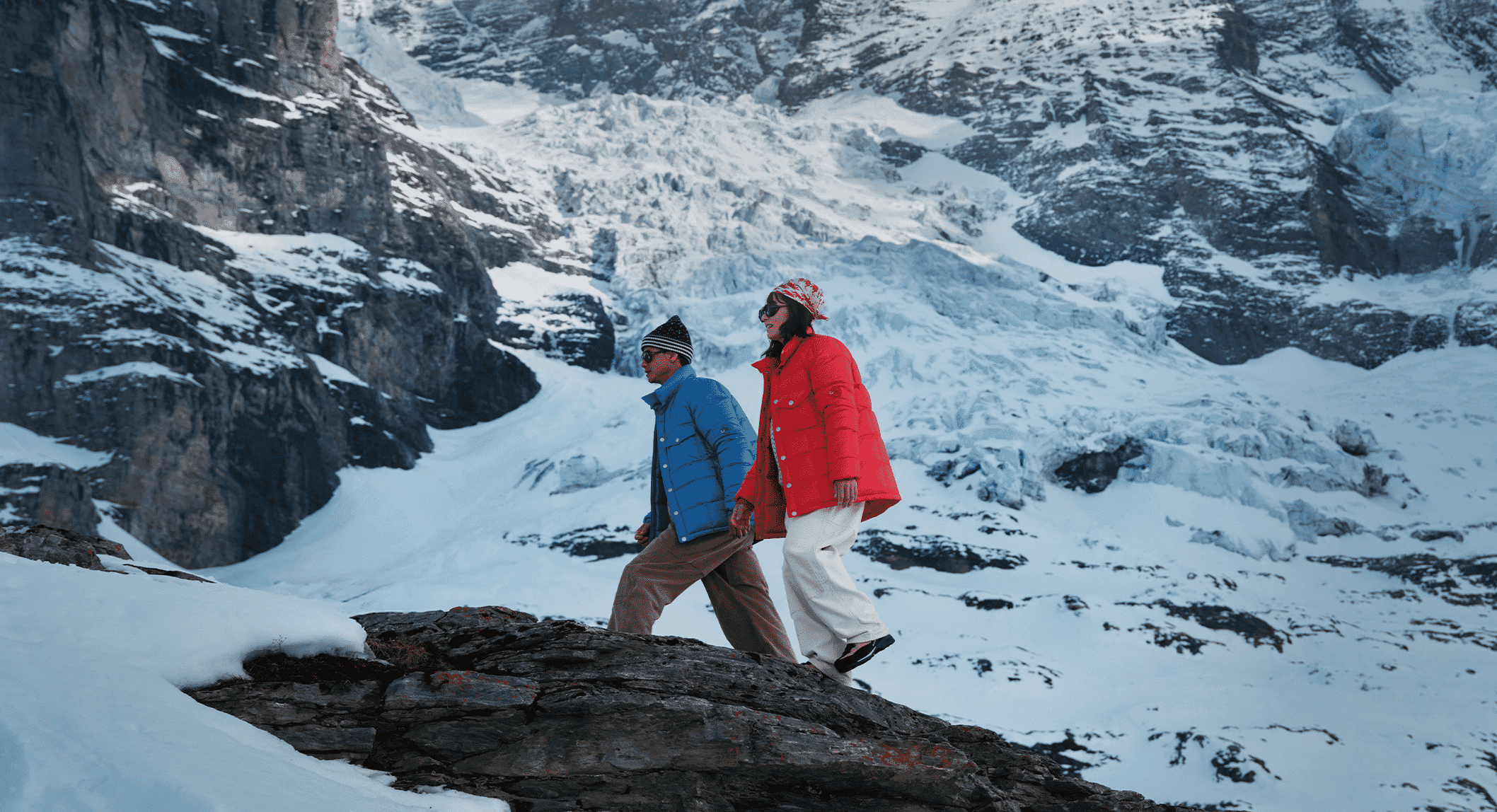 Man and woman walking on a snowy mountain ridge in colorful blue and red oversized puffer jackets.