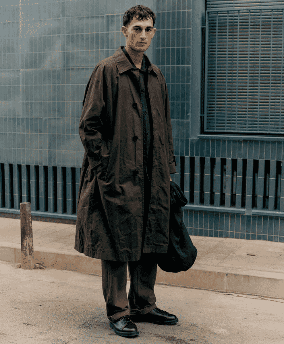 A man in an oversized dark brown waxed cotton trench coat walking on an urban sidewalk.