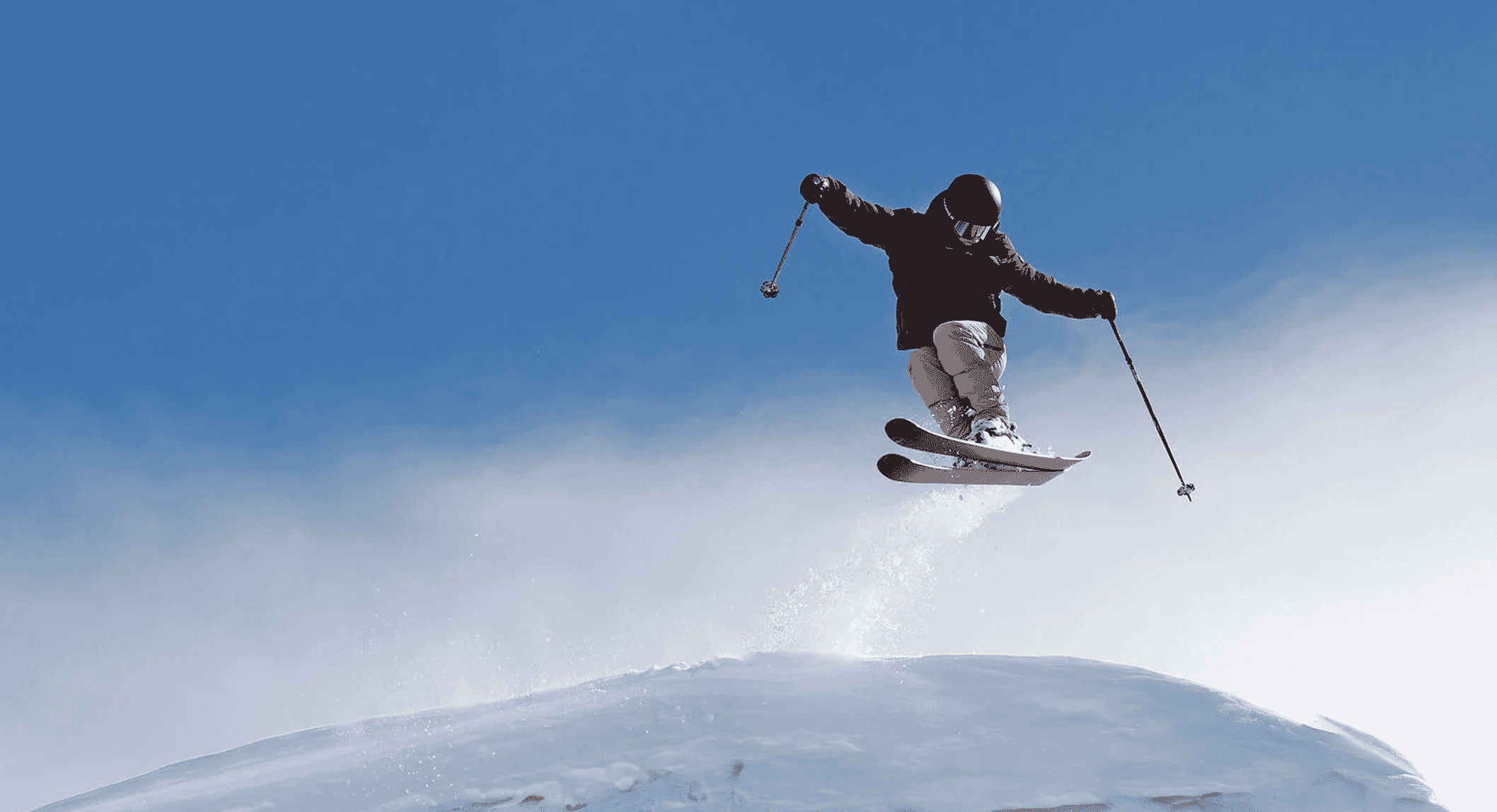 Action shot of a skier in black jacket and helmet performing a jump with snow spray against a blue sky.