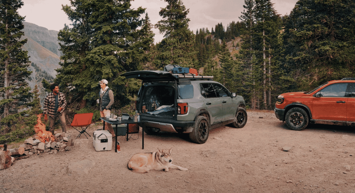 Two men in plaid shirts and vests camping near a fire beside SUVs in a forest.