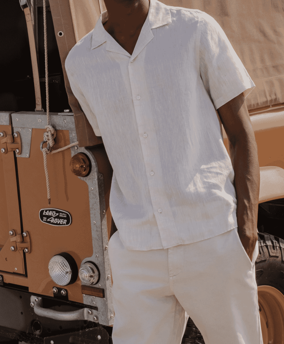 Man wearing a white linen short-sleeve shirt leaning against a vintage off-road vehicle in the sun.