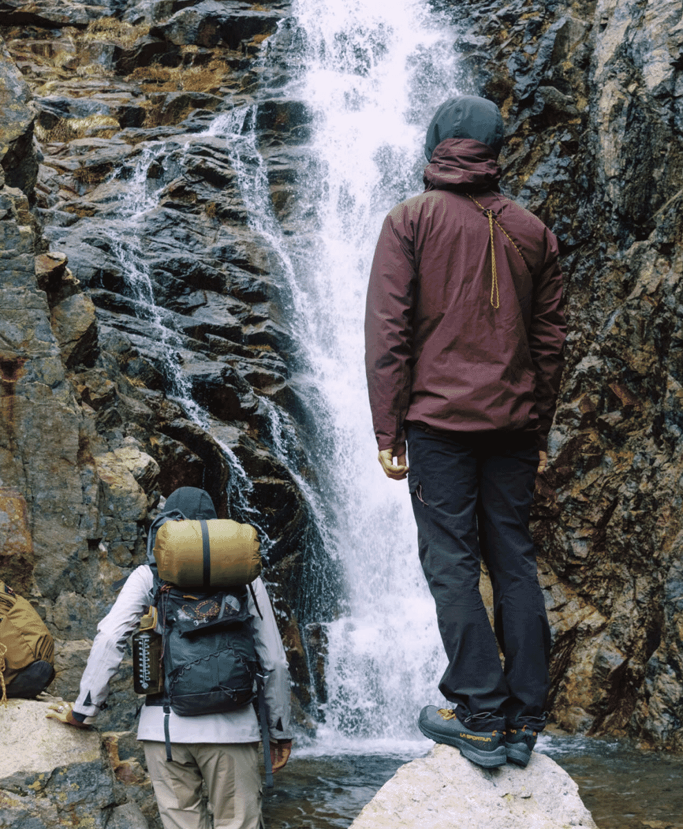 A hiker wearing a maroon technical jacket and black trekking pants standing on a rocky outcrop near a mountain waterfall.