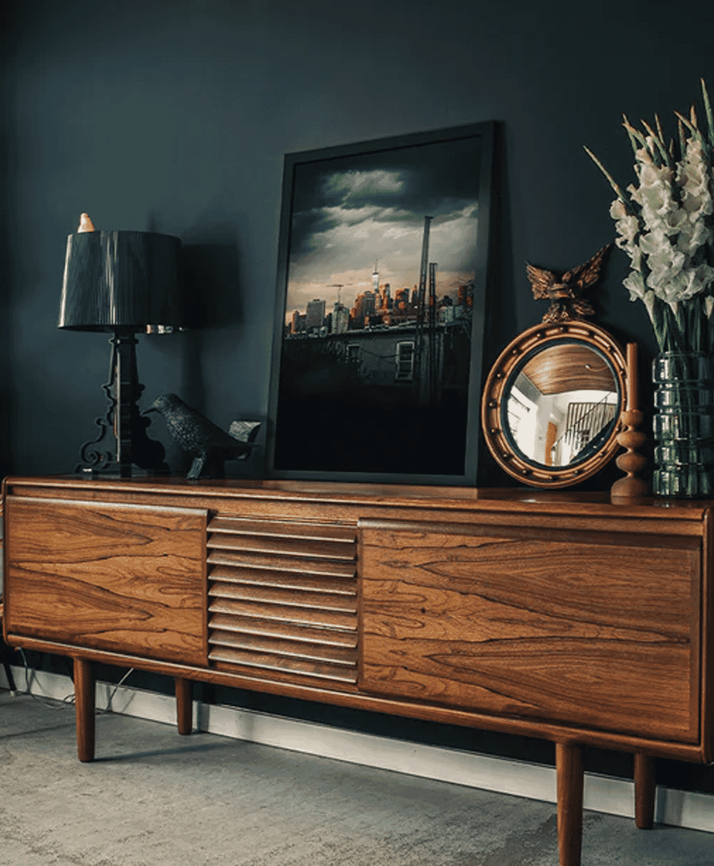 A wooden mid-century credenza featuring a framed urban skyline photograph, a circular mirror, and decorative vases.