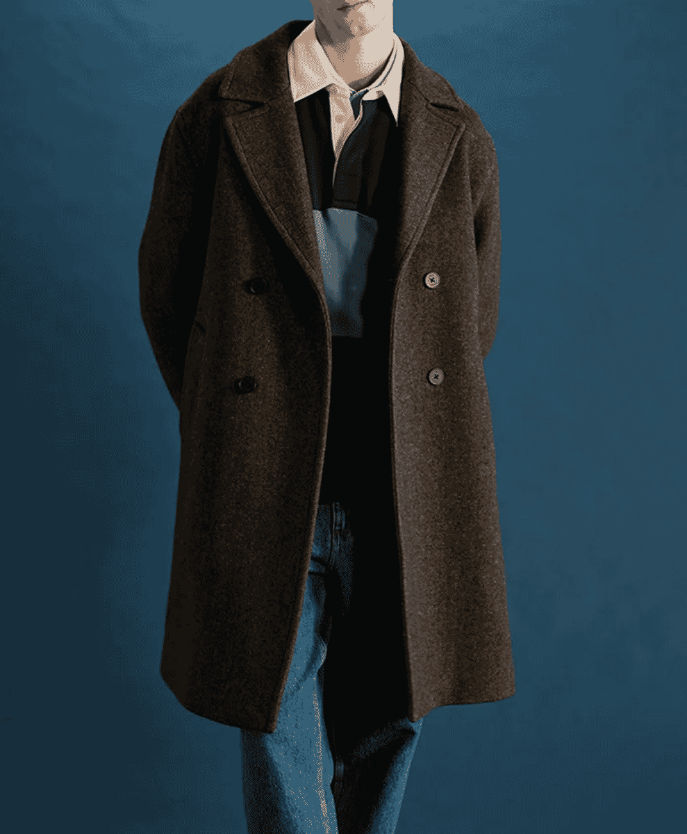 A man in a brown wool coat layered with a polo shirt and sweater against a teal background.