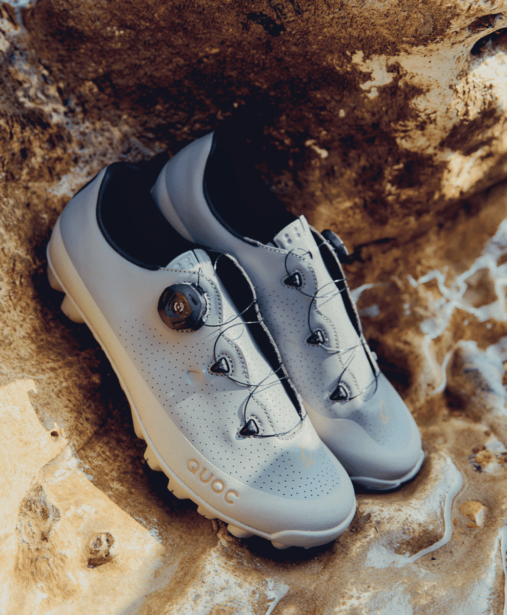QUOC white performance cycling shoes displayed on a textured stone surface.