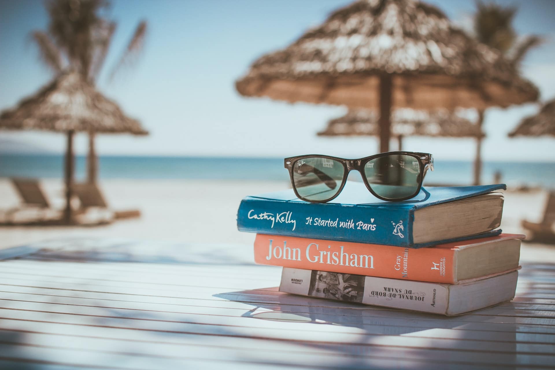 Sunglasses on a stack of books with a blurry background of beach umbrellas and the ocean.