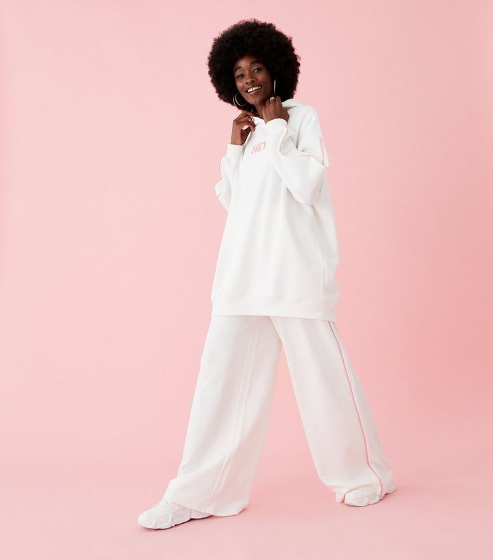 Woman in a white 'HEY' hoodie and matching wide-leg sweatpants against a pink background.