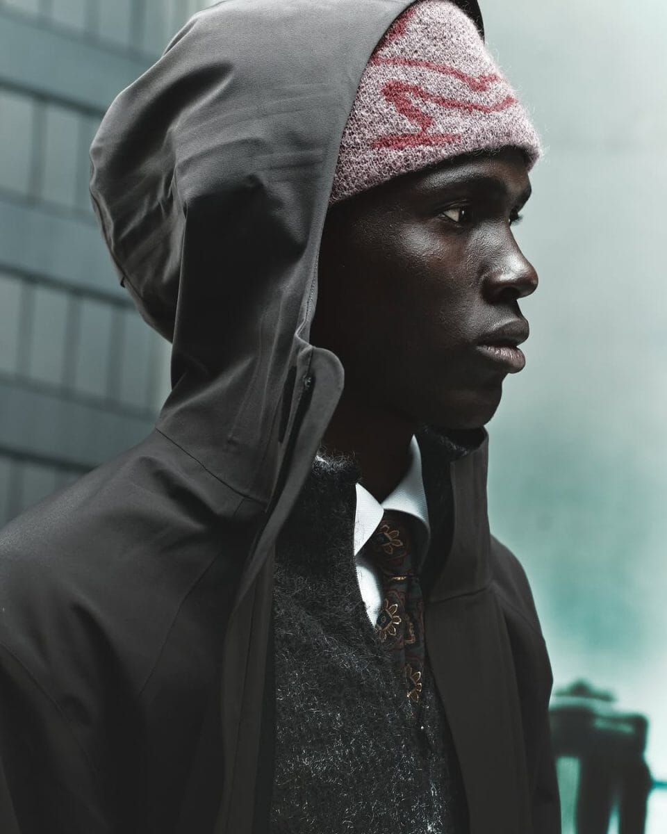 Profile view of a man wearing a dark hooded jacket, a patterned tie, and a wool beanie.