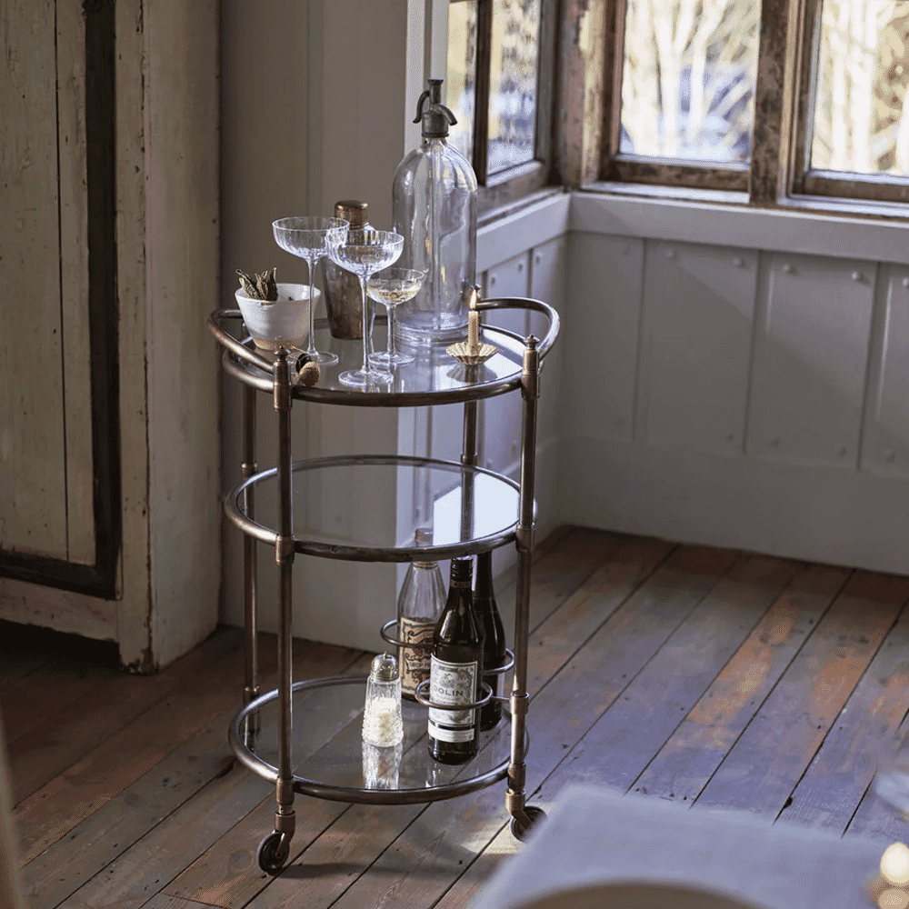 A vintage brass bar cart with glassware in a rustic room with wooden floors and natural light.