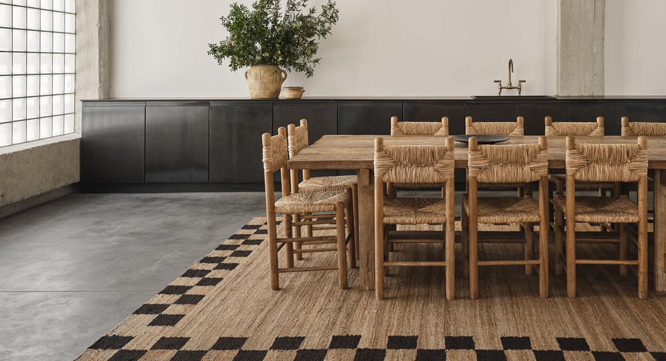 A wide shot of a rustic dining room with a wooden table on a black-and-beige checkered jute rug.
