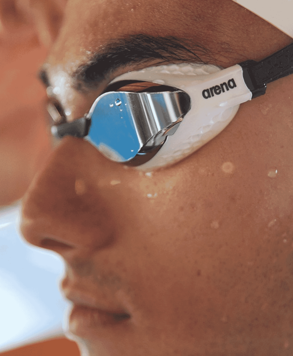 Close-up of a swimmer's face wearing white Arena goggles with reflective lenses and water beads on the skin.
