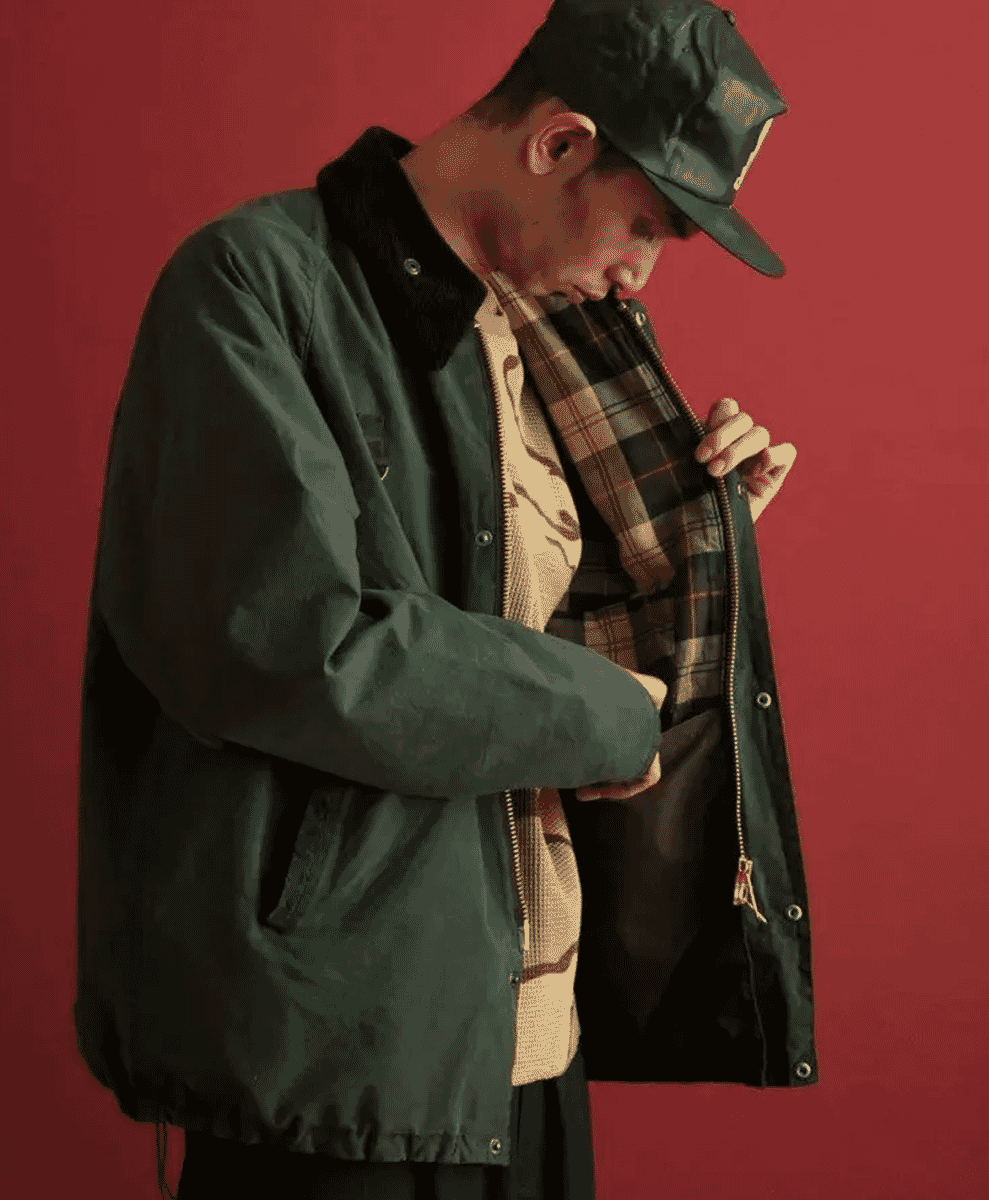 A man in a green waxed jacket with a plaid-lined hood posing against a vibrant red background.