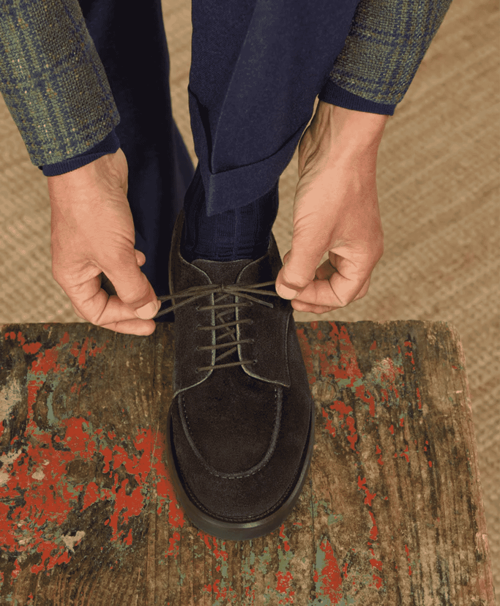 Close-up of a man tying dark suede derby shoes while sitting on a weathered wooden bench.