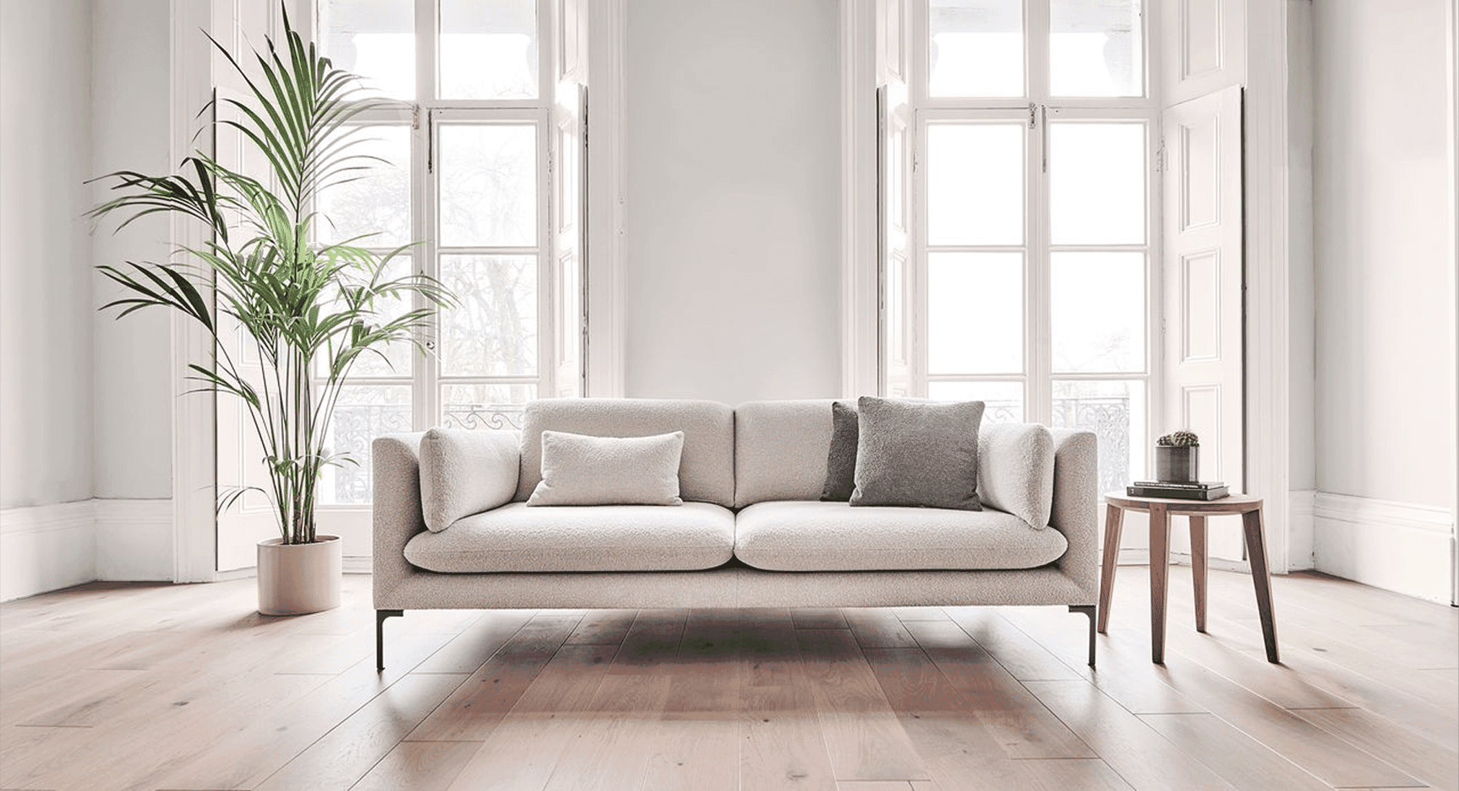 Minimalist living room with a light beige sofa, wooden floors, and large windows allowing natural light.