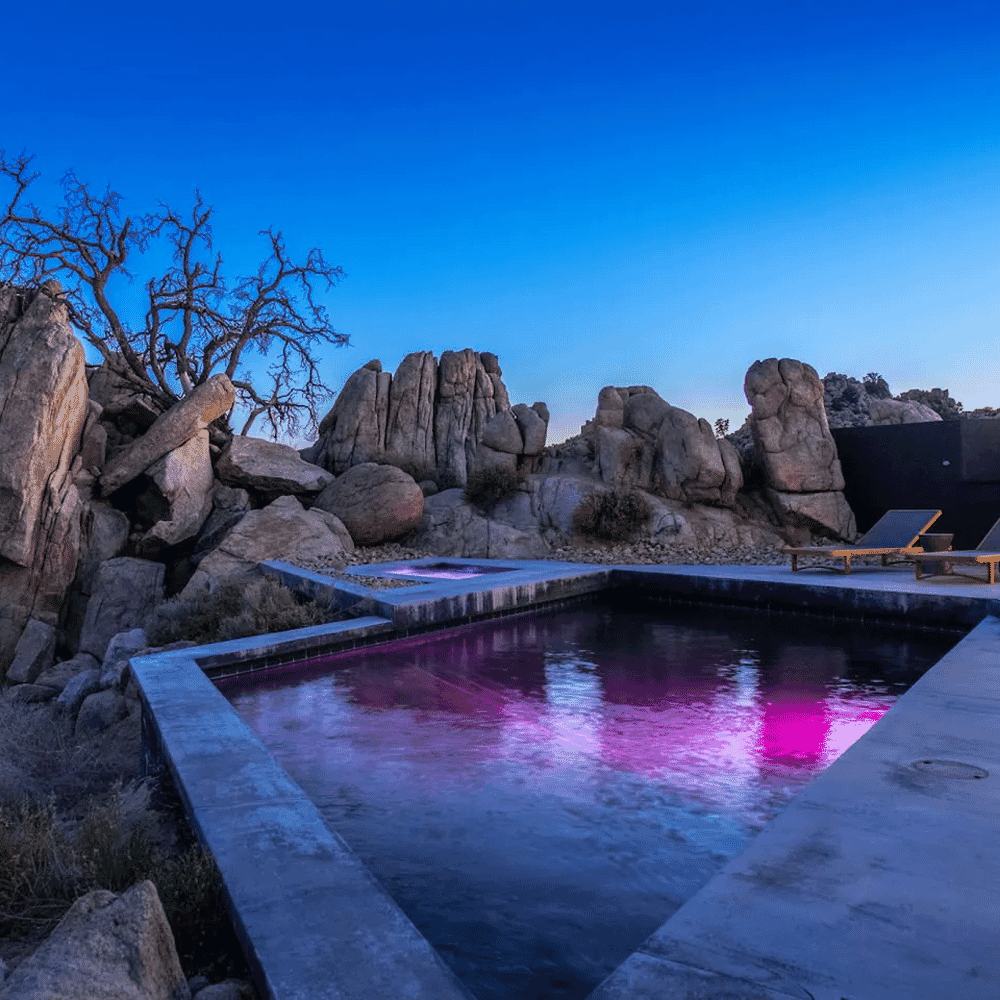 A serene twilight scene featuring a modern pool with purple lighting and minimalist lounge chairs.