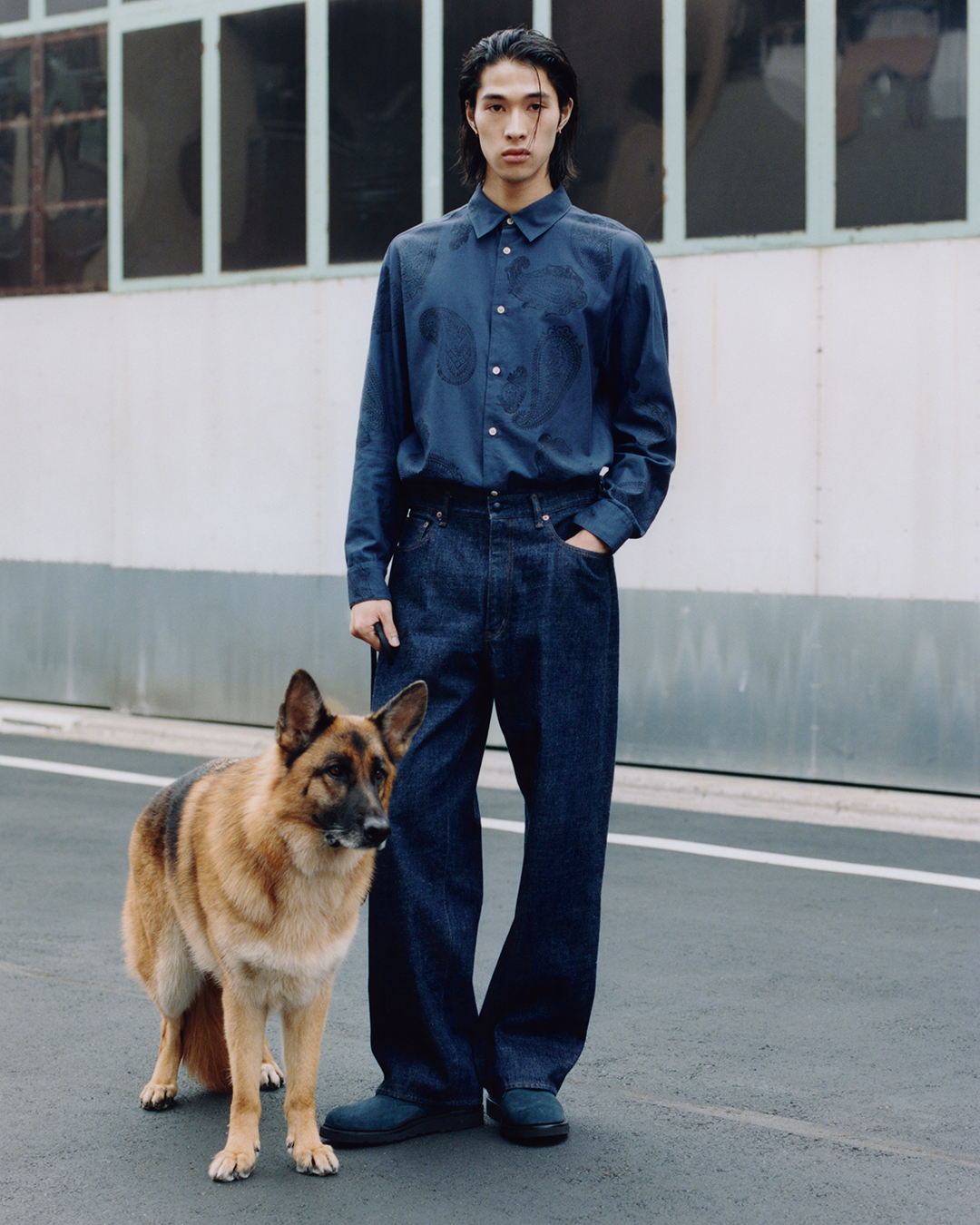 A man in a dark blue paisley shirt and wide-leg denim stands with a German Shepherd on a city street.