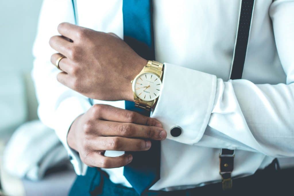 Close-up of a man in formal attire and suspenders adjusting his shirt cuff to reveal a gold Rolex watch.