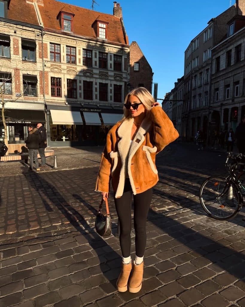 Woman in a tan shearling coat with white fleece lining and platform boots standing on a cobblestone street.