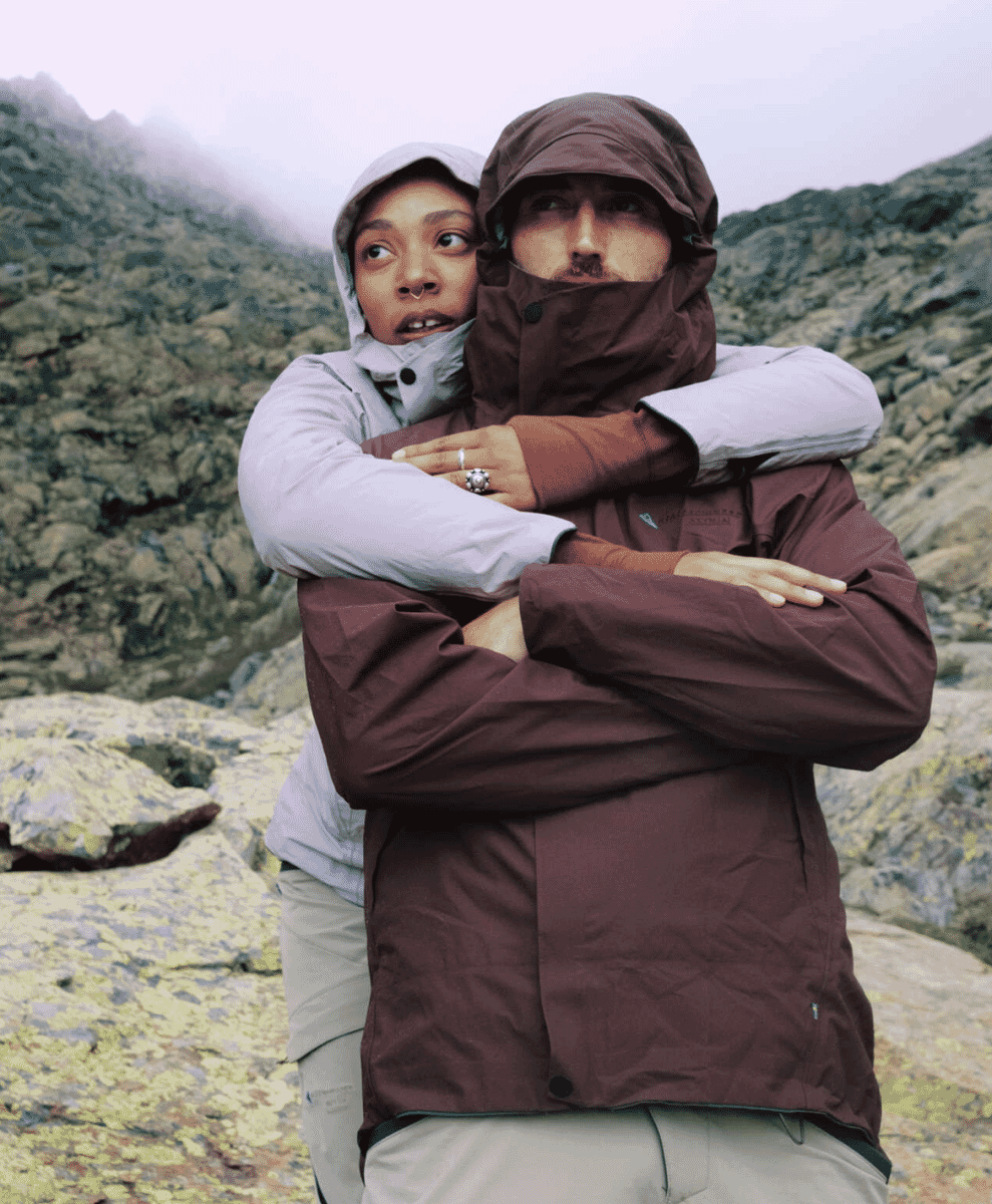 A couple embracing in misty mountain terrain wearing light gray and brown waterproof hooded jackets.