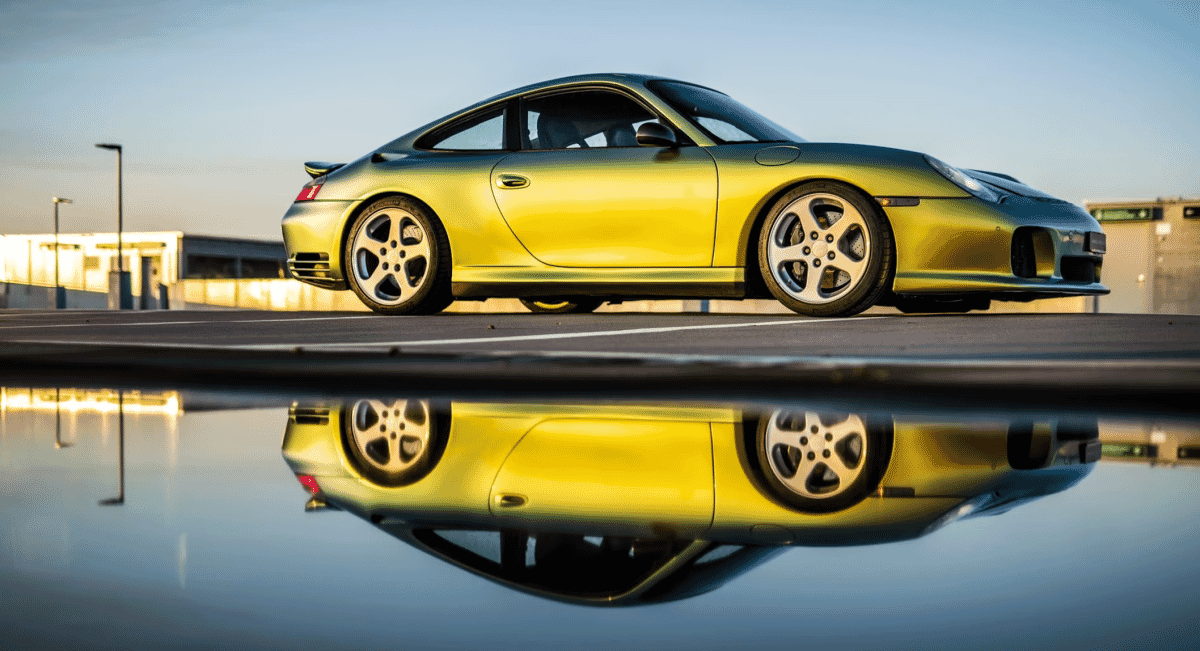 Metallic gold Porsche 911 reflected in a puddle on a wet road at golden hour.
