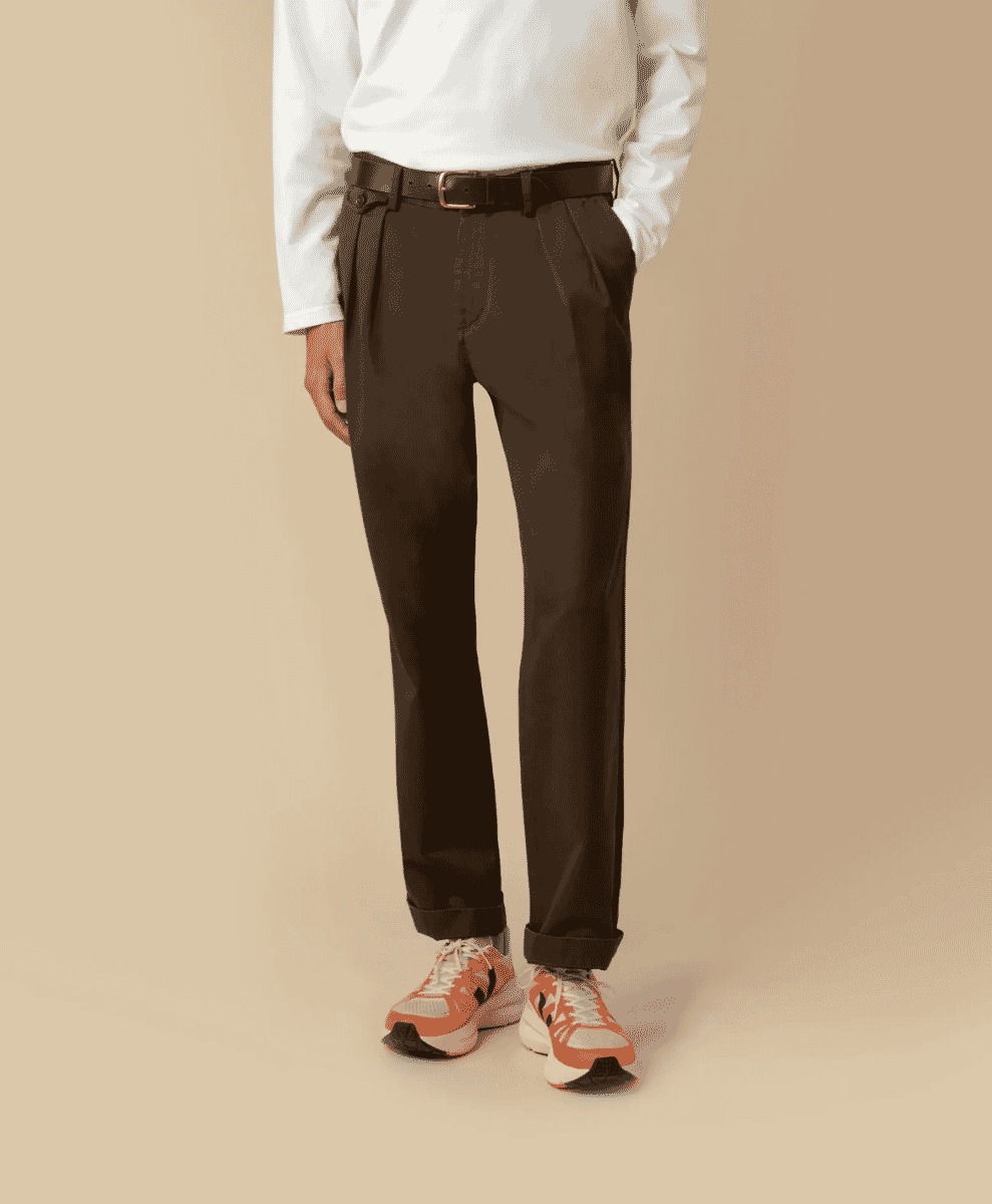 A man models dark brown pleated trousers with a white shirt and vibrant orange sneakers.