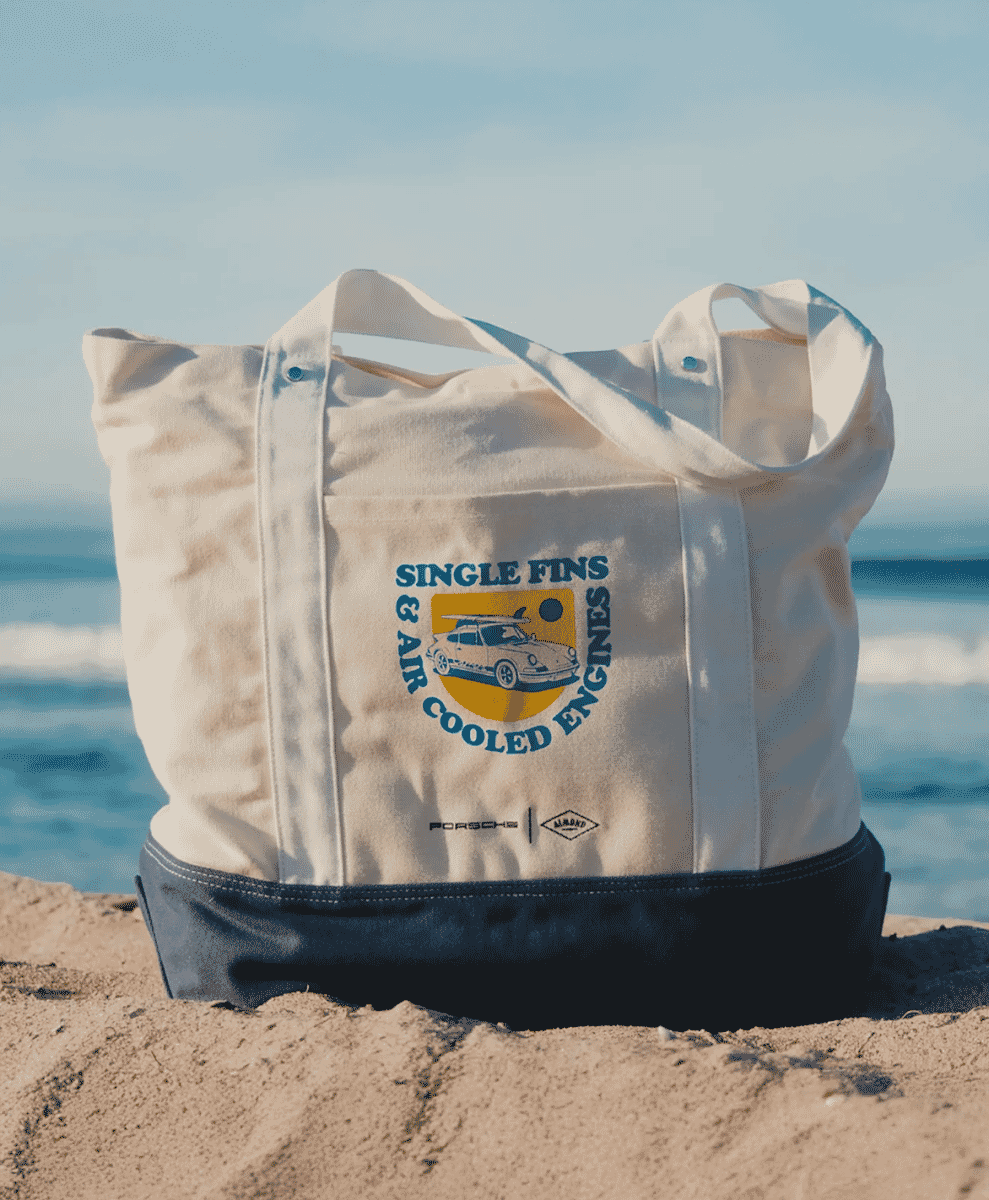 A white canvas tote bag with surf-themed graphic on a sandy beach.