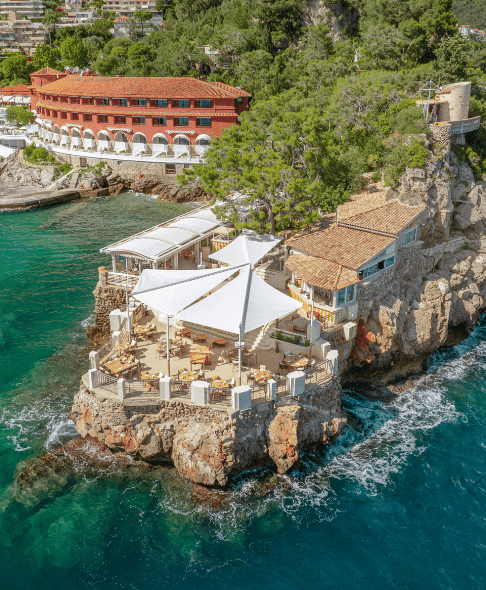 Aerial view of a luxury cliffside restaurant terrace with white umbrellas overlooking the sea.