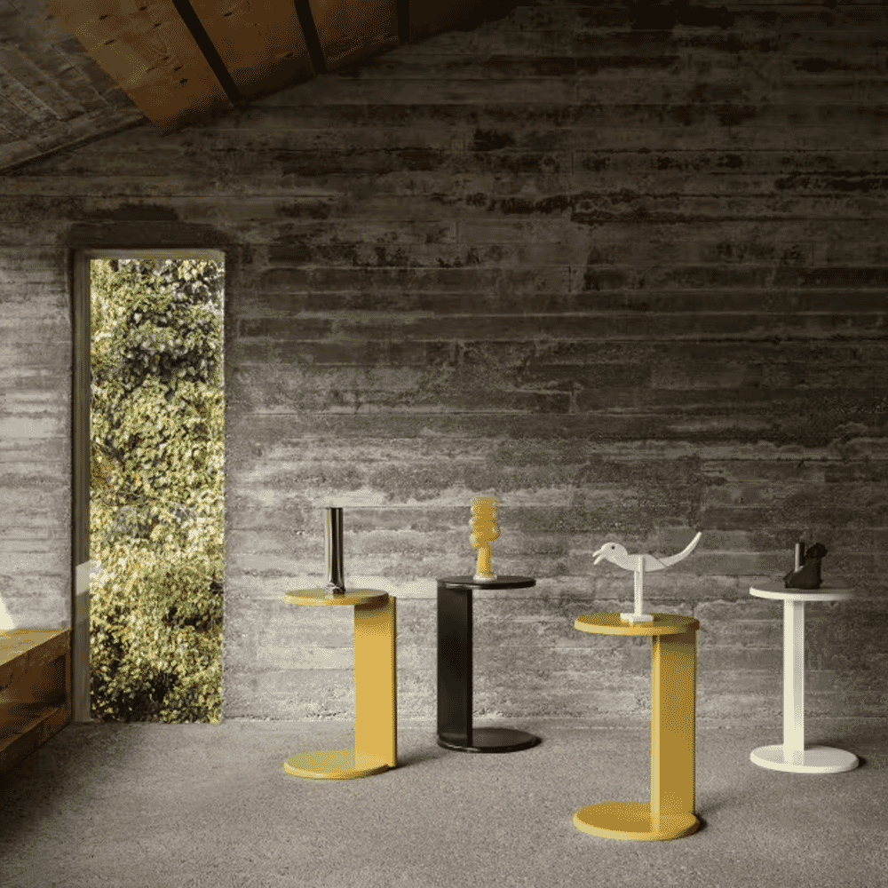 Four minimalist side tables in yellow, black, and white set against an industrial concrete interior.