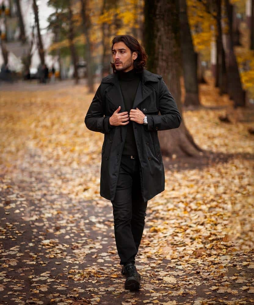 Man wearing a long black trench coat walking through an autumn park with golden leaves.