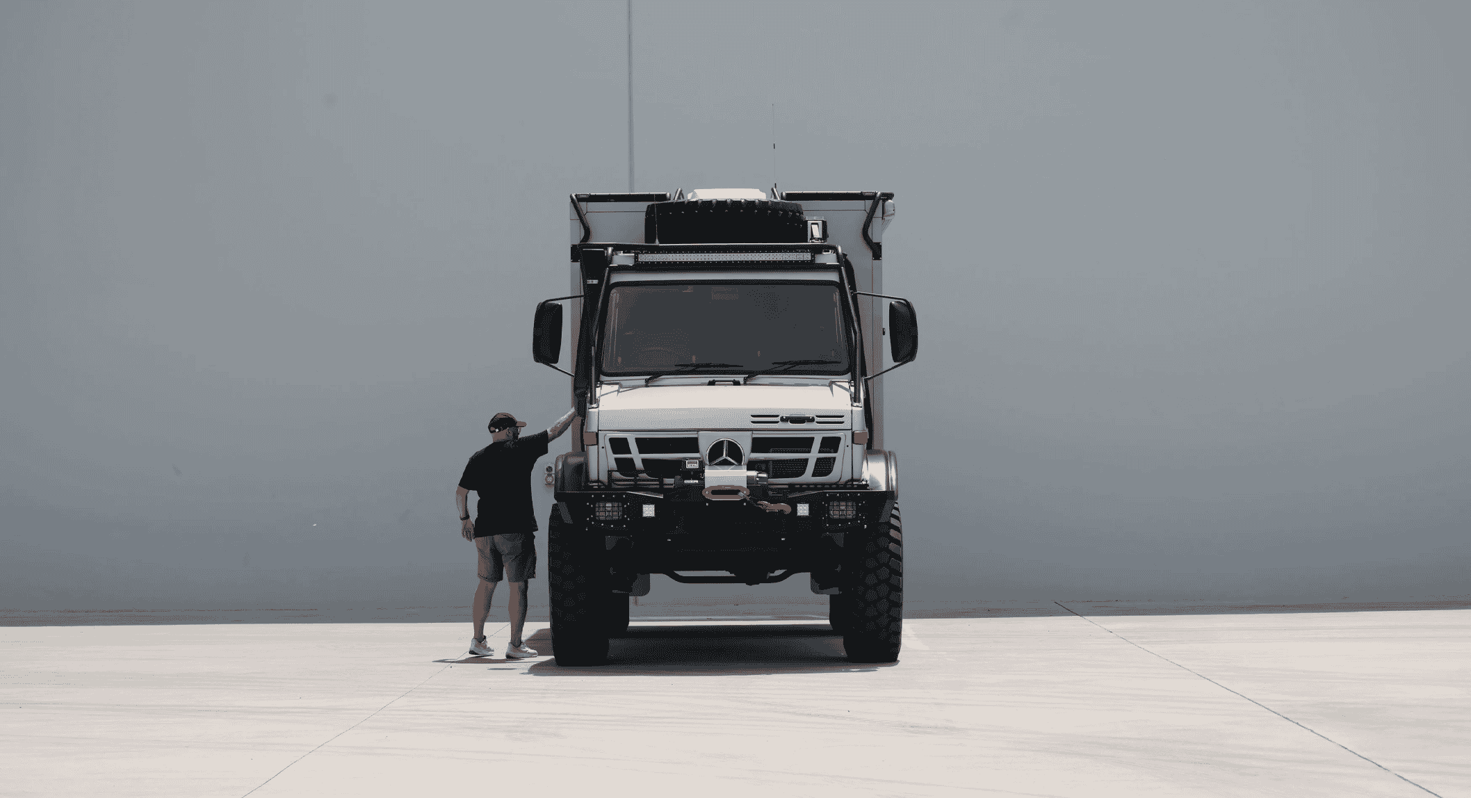 A man in casual outdoor gear stands next to a massive white Mercedes-Benz Unimog in a minimalist warehouse setting.